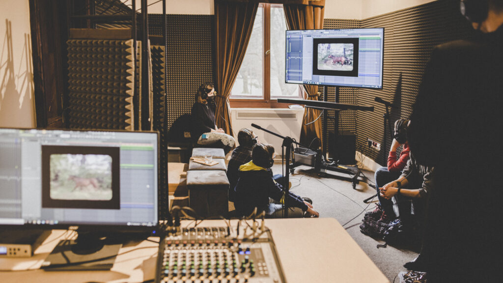 Studio nagraniowe. Na pierwszym planie ekran monitora i panel sterowania dźwiękiem. Na drugim planie pufach i podłodzie siedzi kilka osób, wpatrujących się w ekran telewizora.