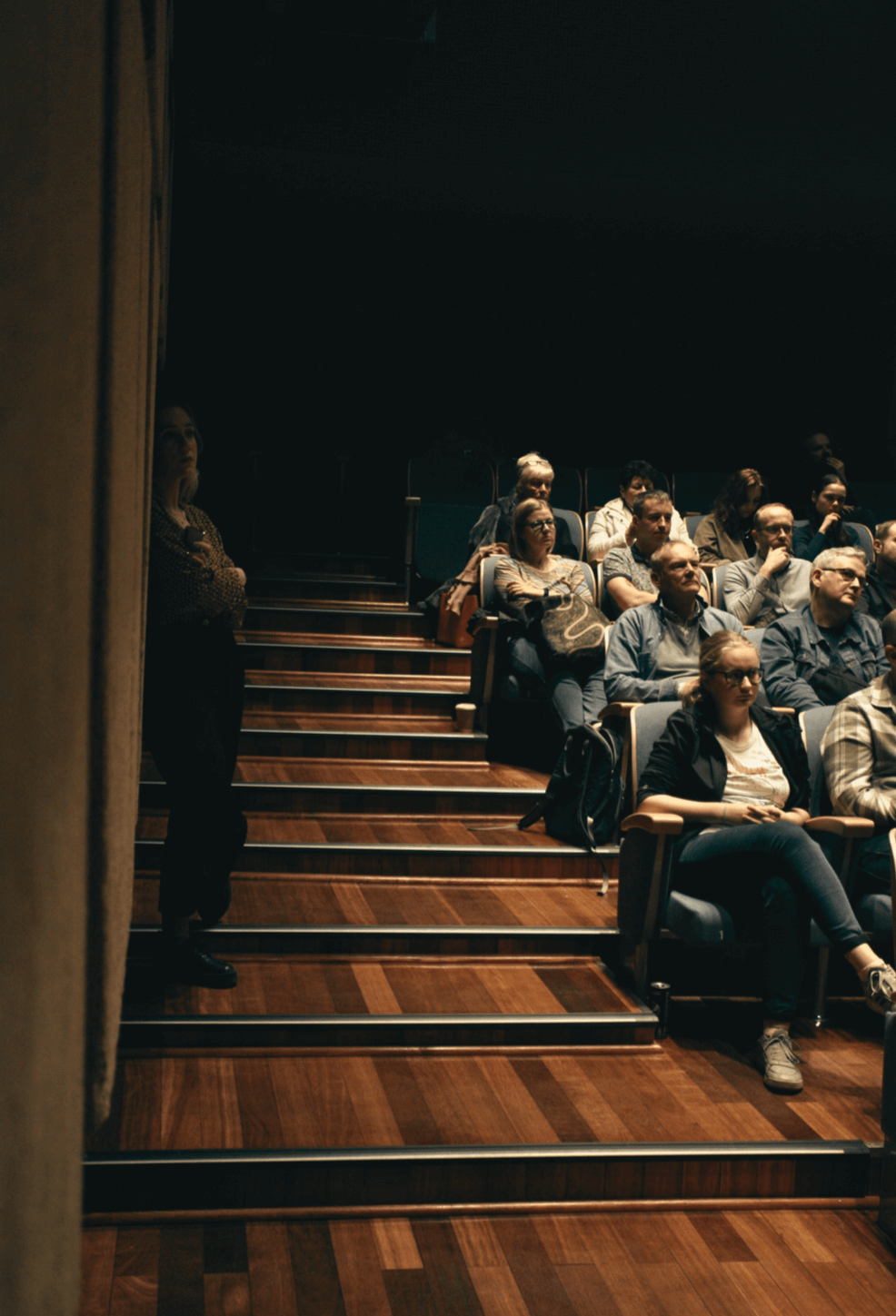 Sala kinowa. Widzowie siedzą w fotelach. Na schodach obok foteli stoi kobieta.