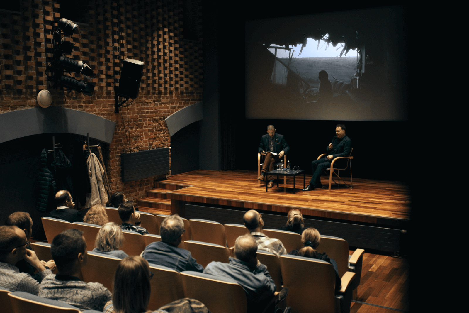 Sala kinowa. Od tyłu widoczni widzowie, siedzący w fotelach. Przed nimi scena oraz ekran projekcyjny. Na scenie, dwa krzesełka, w których siedzi dwóch mężczyzn.