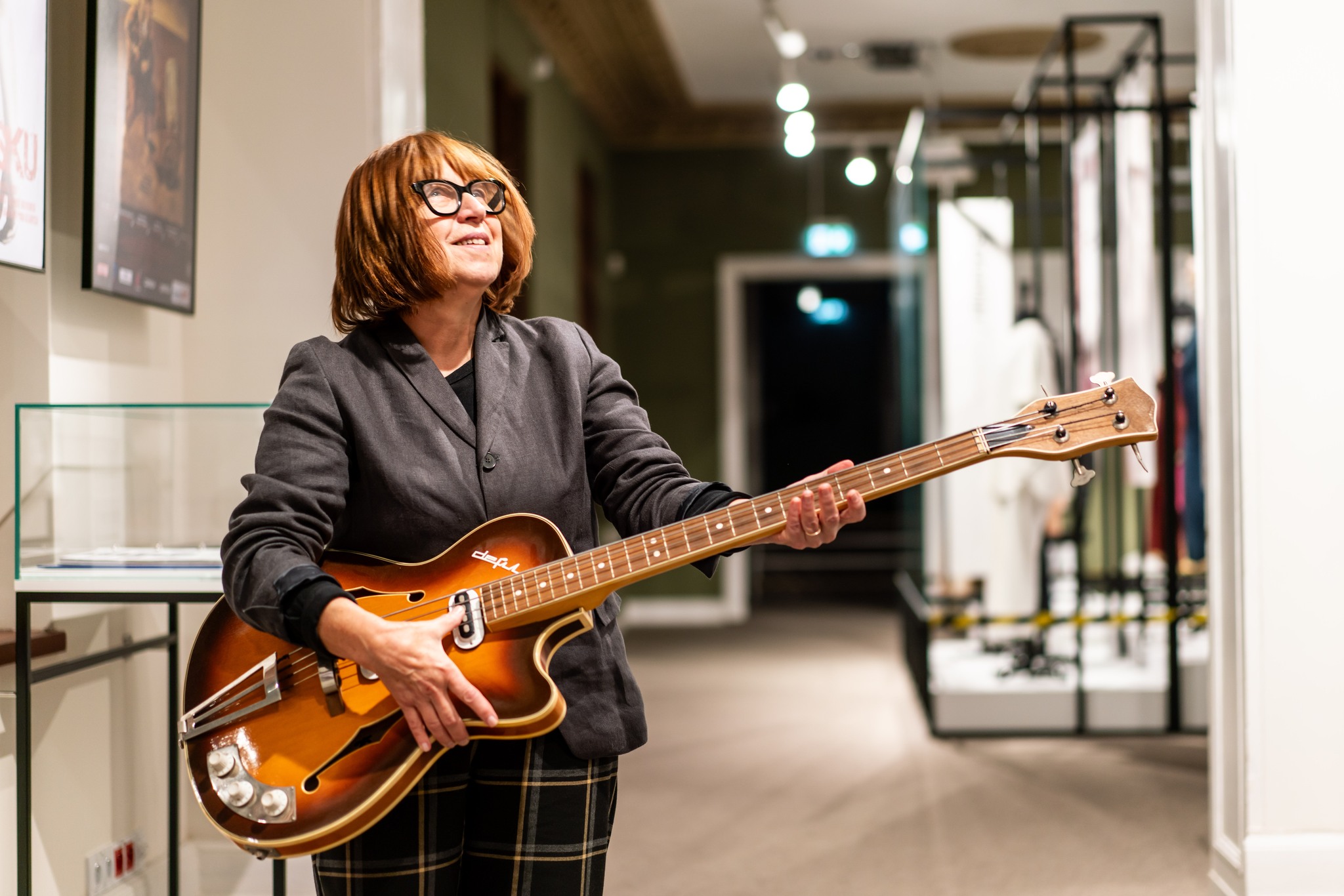 Kobieta w średnim wieku z elektryczną gitarą. W tle muzeum i ekspozycje.