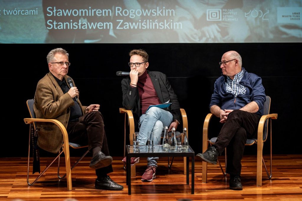 Trzej mężczyźni siedzący na krzesłach na scenie. Przed nimi stolik. Za nimi ekran.