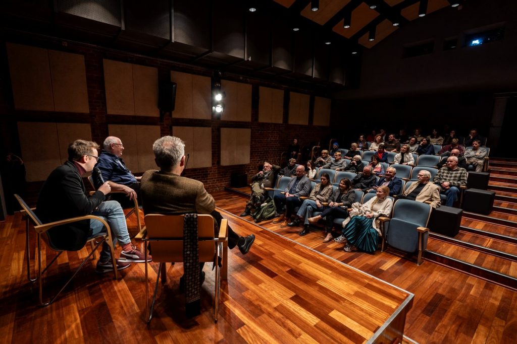 Sala kinowa. Ludzie siedzący w fotelach na widowni patrzą w stronę sceny. Na scenie od tyłu ukazano trzech mężczyzn siedzących na krzesłach. Mężczyzna po środku trzyma przy ustach mikrofon.