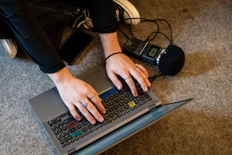 Osoba siedząca po turecku na podłodze, widoczna tylko kawałkiem. Ręce trzyma na klawiaturze laptopa przed sobą. Obok laptopa leży dyktafon z mikrofonem. W prawym górnym rogu rożowa myszka komputerowa.