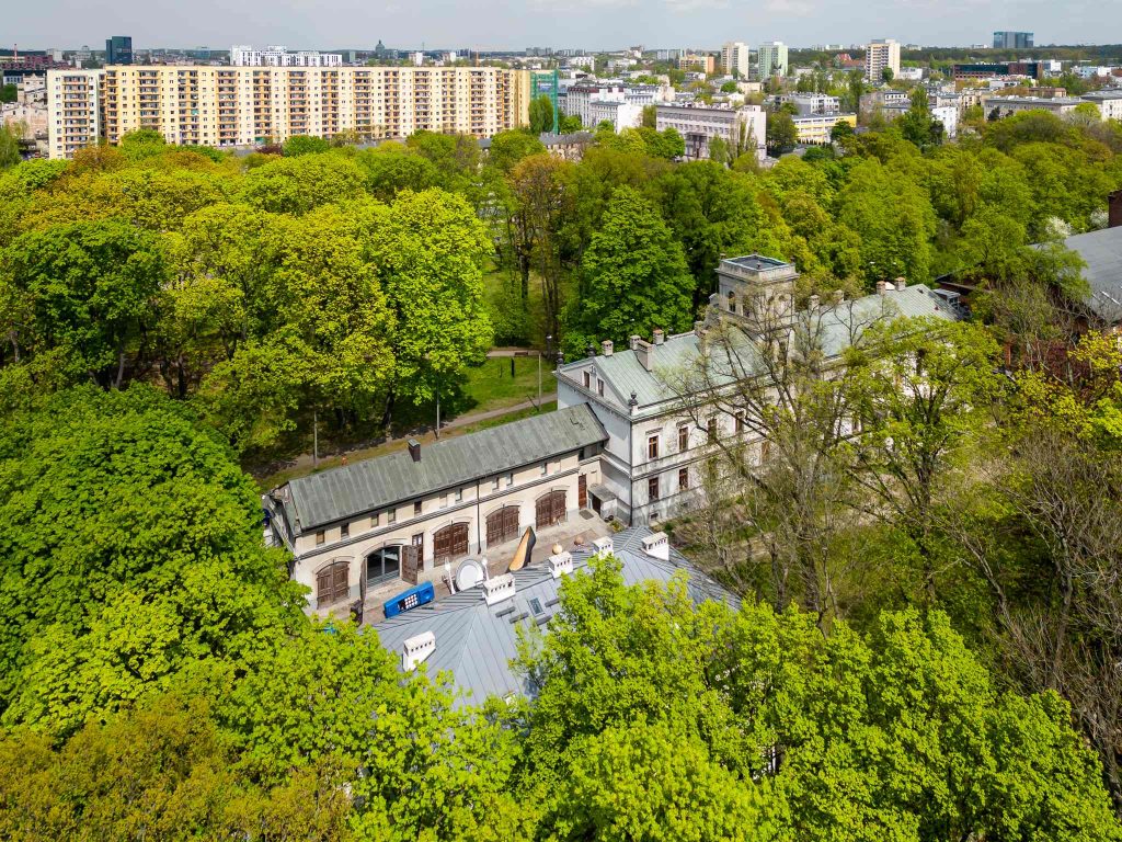 Budynek Muzeum Kinematografii w Łodzi wiosną, widok z lotu ptaka. Miejsce to otoczone jest zielonymi drzewami.