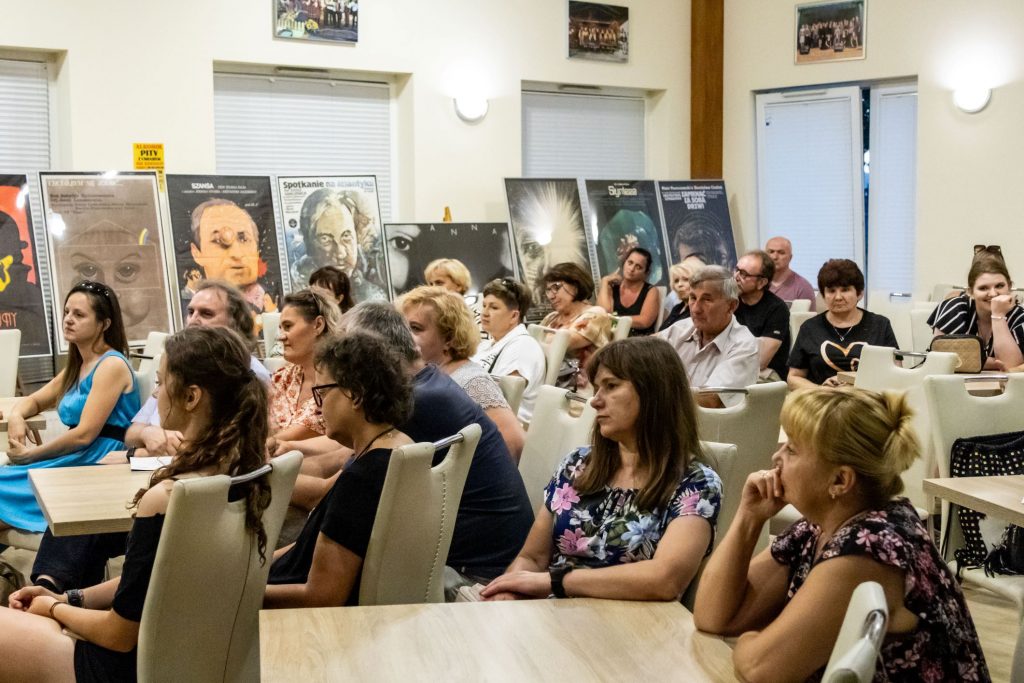 Grupa kilkunastu osób siedzących w sali przy stolikach. W tle pod ścianą wystawa różnych plakatów filmowych. Plakaty stoją na sztalugach. Za plakatami duże, zasłonięte roletami okna.