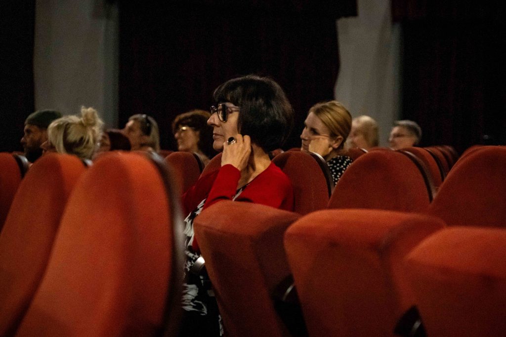 Ludzie siedzący w kinowych fotelach, skupieni na wydarzeniu przed nimi. Kobieta w czerwonym swetrze na pierwszym planie spogląda w stronę ekranu lub sceny, podpierając twarz dłonią.