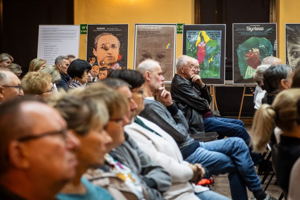 Widok na publiczność siedzącą w rzędach, skierowaną w stronę prezentacji. Na ścianie za nimi wiszą plakaty z różnorodnymi ilustracjami.