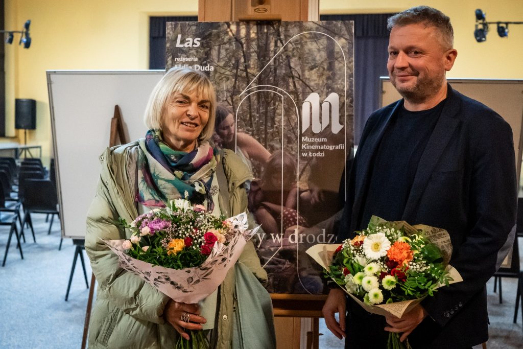 Kobieta z jasnymi włosami trzyma bukiet kwiatów i stoi obok mężczyzny, który także trzyma kwiaty. Za nimi znajduje się plakat promujący wydarzenie.