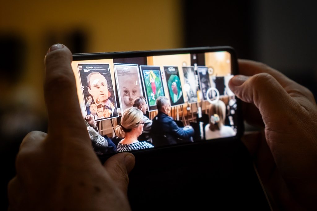 Kadr zbliżony na ręce trzymające telefon, który robi zdjęcie plakatów filmowych i publiczności siedzącej w rzędach. Na ekranie telefonu widoczne są te same plakaty i ludzie.