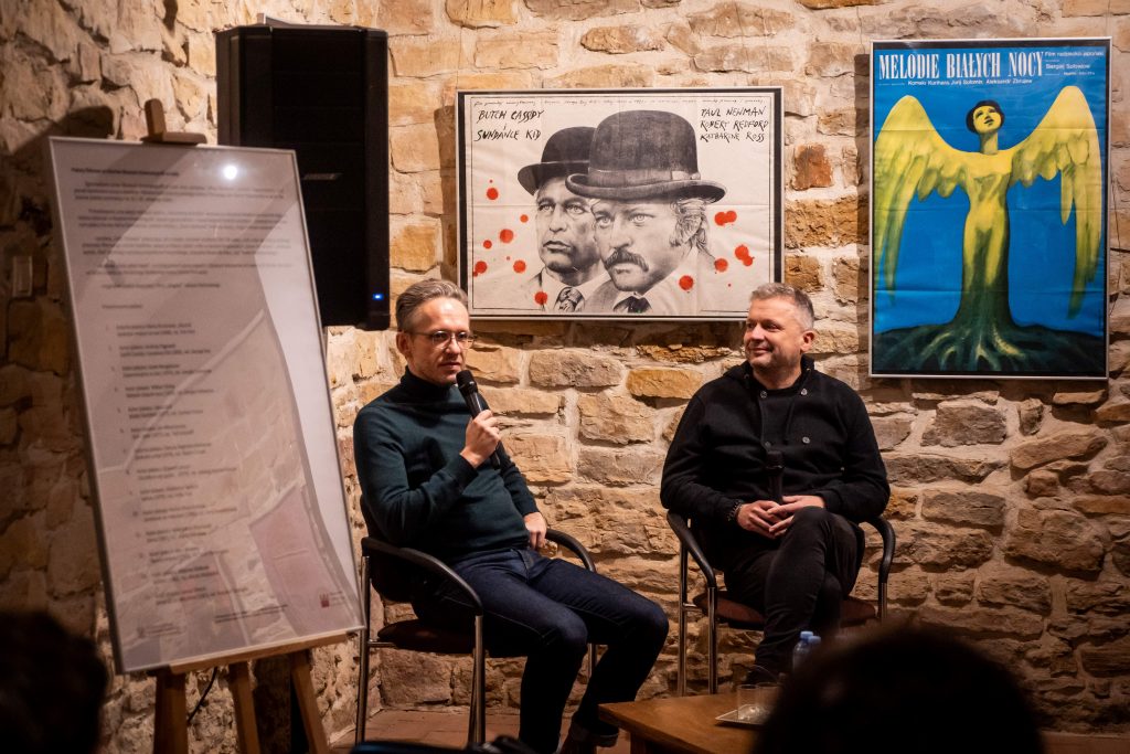 Dwóch mężczyzn siedzi na scenie, jeden z nich trzyma mikrofon. Za nimi widać plakaty filmowe na tle kamiennej ściany, w tym plakat do filmu "Butch Cassidy i Sundance Kid".