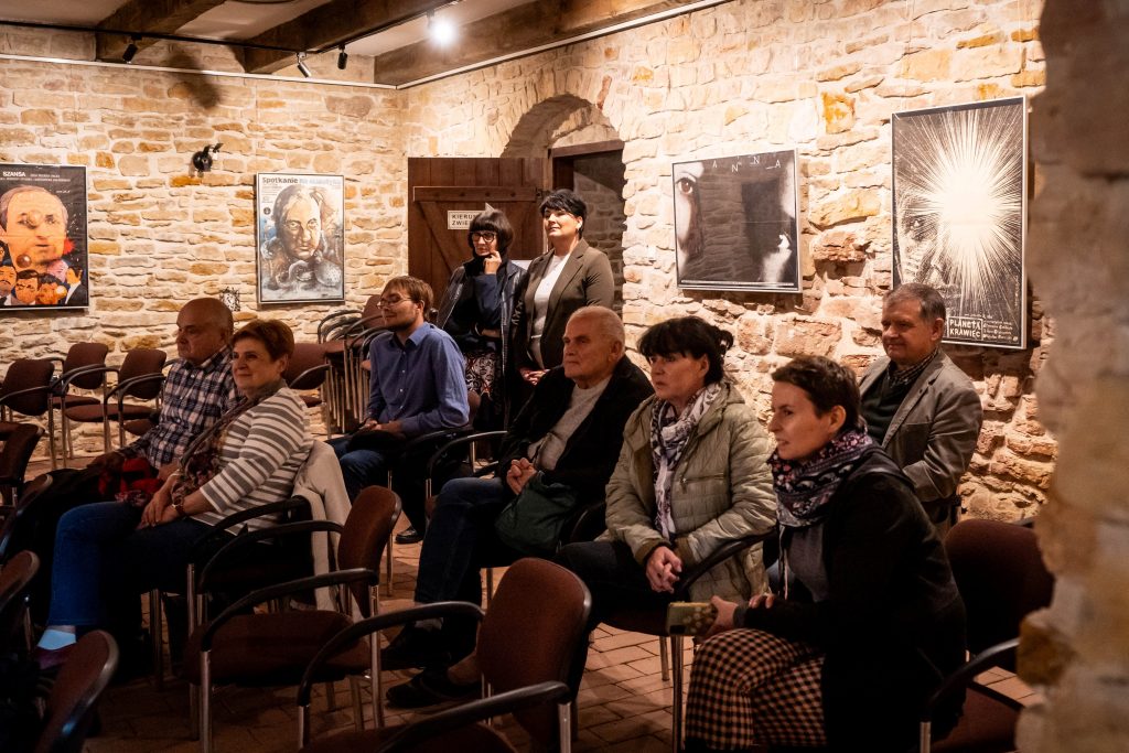 Grupa osób siedzi w sali z kamiennymi ścianami. Na ścianach widoczne są plakaty filmowe. Wszyscy patrzą w jednym kierunku, sugerując, że słuchają prelekcji.